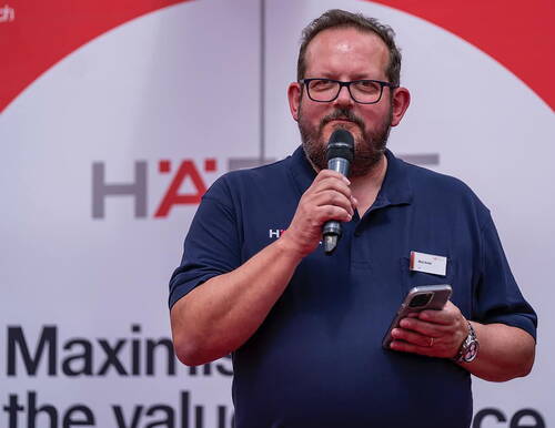 Mittelalterlicher Mann mit Brille hält ein Mikrofon und ein Smartphone, während er vor einem rot‑weißen Konferenzbanner spricht
