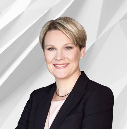 Lächelnde Geschäftsfrau mit kurzem blonden Haar im dunklen Blazer vor einem abstrakten, hellgrauen Hintergrund