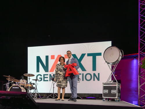Zwei lächelnde Personen stehen auf einer Bühne vor einer großen „NEXT GENERATION“-Leinwand und halten gemeinsam ein rotes A, umgeben von Schlagzeug und Veranstaltungstechnik.