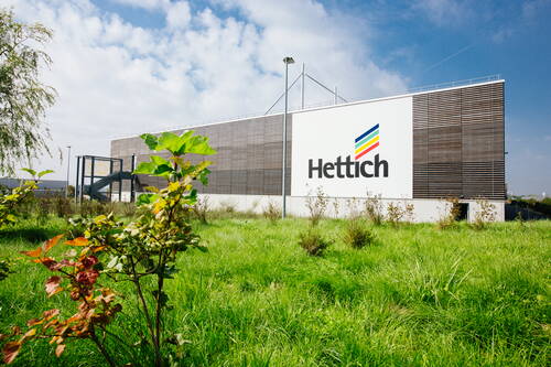 Modernes Firmengebäude von Hettich mit großer Logo­fassade hinter einer grünen, naturbelassenen Wiese unter blauem Himmel