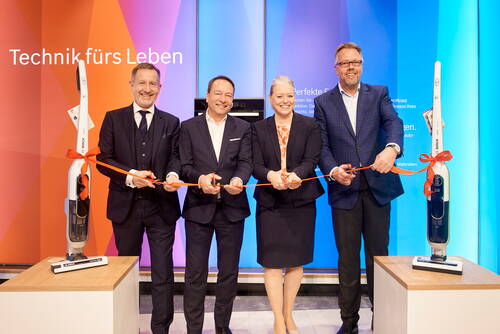 Vier Geschäftsleute in Anzügen durchschneiden gemeinsam ein rotes Band zwischen zwei Bosch-Staubsaugern vor einer farbigen „Technik fürs Leben“-Wand bei einer Produkteinführung.