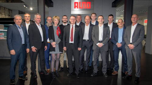 Gruppe von Geschäftsleuten in Anzügen und Businesskleidung posiert lächelnd für ein Gruppenfoto in einem modernen Firmenshowroom mit leuchtendem ALNO-Logo im Hintergrund