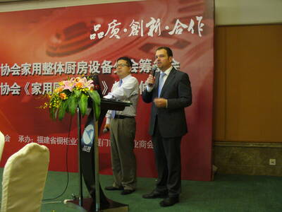 Zwei Männer halten eine Präsentation auf einer Konferenzbühne vor rotem Hintergrund mit chinesischer Schrift, während einer am Rednerpult mit Blumenarrangement steht und der andere mit Mikrofon spricht.