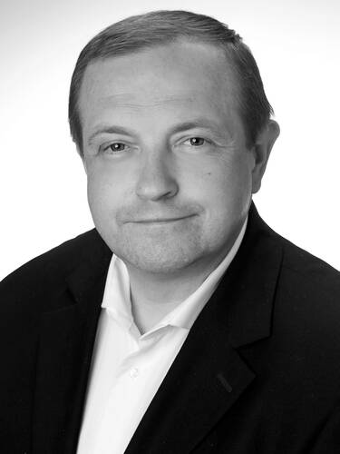 Mittelalterlicher Mann im Anzug mit weißem Hemd lächelt leicht in die Kamera vor neutralem Hintergrund.
