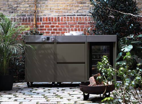 Moderne Outdoor-Küchenzeile aus Metall mit integriertem Kühlschrank und Feuerschale auf einem gepflasterten Innenhof vor einer Backsteinwand, umgeben von üppigen Pflanzen