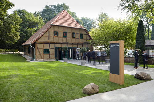 Historisches Fachwerkhaus mit rotem Ziegeldach, gepflegter Rasenfläche und elegant gekleideten Menschen bei einer Veranstaltung neben einer modernen Holzstele mit Firmenlogo.
