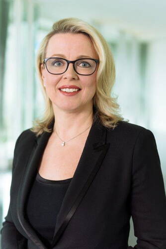 Blonde Frau mit Brille und schwarzem Blazer lächelt freundlich in moderner, hell beleuchteter Büro-Umgebung