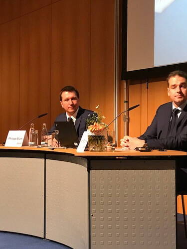 Zwei Männer im Anzug sitzen an einem Konferenztisch mit Namensschildern, Mikrofonen und Blumenarrangement und bereiten sich auf eine Podiumsdiskussion vor.