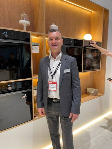 Geschäftsmann im Anzug präsentiert moderne Einbauküchengeräte in einem hell beleuchteten Messestand aus Holz.