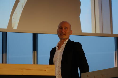 Glatzköpfiger Mann im Anzug hält einen Vortrag an einem Rednerpult vor einer großen Projektionsleinwand in einem modernen Konferenzraum