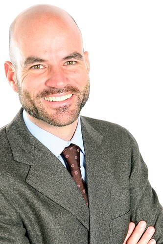 Glatzköpfiger Mann im Anzug mit verschränkten Armen lächelt selbstbewusst vor weißem Hintergrund