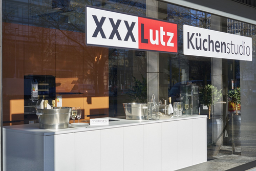 Modernes Küchenstudio-Schaufenster von XXXLutz mit weißer Küchenzeile, Champagnerkühlern und Gläsern in heller Straßenlage
