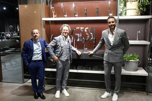 Drei elegant gekleidete Männer posieren vor einer Ausstellungswand mit modernen Wasserhähnen und großem Gessi‑Logo in einem Showroom.
