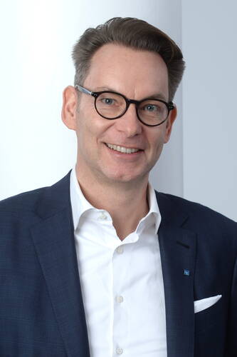 Lächelnder Mann mit Brille in dunkelblauem Anzug und weißem Hemd vor hellem Hintergrund