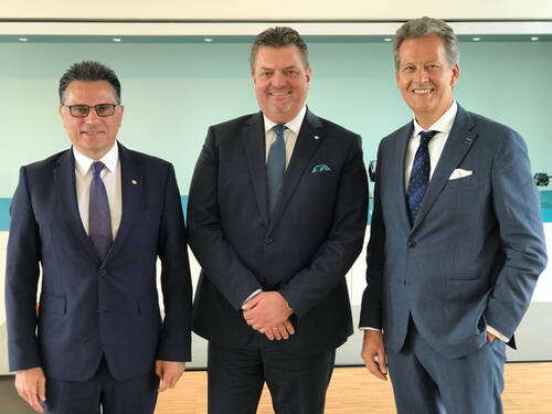 Drei lächelnde Geschäftsleute in Anzügen stehen nebeneinander in einem modernen, hellen Konferenzraum