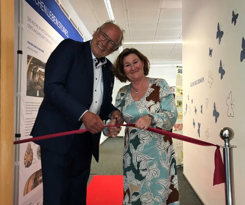 Ein lächelnder Mann im Anzug und eine Frau im gemusterten Kleid durchschneiden gemeinsam ein rotes Band zur feierlichen Eröffnung eines Ausstellungsbereichs mit Infotafeln und Schmetterlingsdekoration an der Wand.