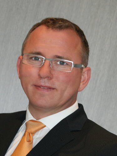 Mittelälterlicher Mann mit Brille im dunklen Nadelstreifenanzug und orangefarbener Krawatte vor neutral grauem Hintergrund, der freundlich in die Kamera blickt.