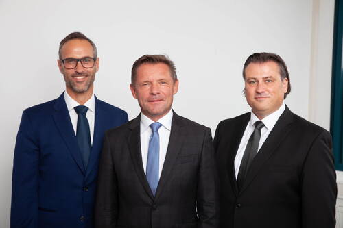 Drei lächelnde Geschäftsleute in Anzügen stehen nebeneinander vor neutralem Hintergrund und blicken direkt in die Kamera