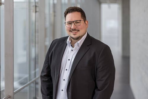 Mann mit Brille im dunklen Anzug und weißem Hemd lächelt in einem modernen, lichtdurchfluteten Büroflur in die Kamera