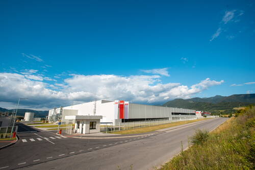 Großer moderner Industriekomplex mit Zufahrtsstraße und Sicherheitsgebäude vor einer Bergkulisse unter klarem blauem Himmel