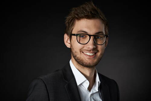 Junger Mann mit Brille und Bart lächelt im dunklen Anzug vor schwarzem Hintergrund