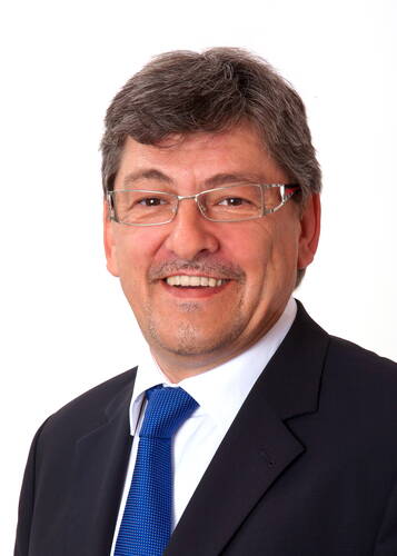 Lächelnder Mann mittleren Alters mit Brille im dunklen Anzug und blauer Krawatte vor weißem Hintergrund