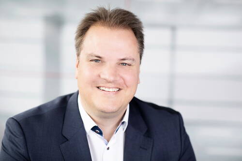 Lächelnder Mann im Business-Anzug blickt direkt in die Kamera vor unscharfem, hellen Bürohintergrund