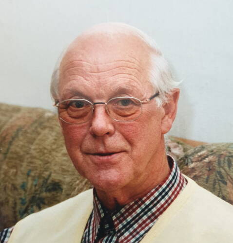 Älterer Mann mit Brille und kariertem Hemd sitzt lächelnd auf einem gemusterten Sofa vor hellem Hintergrund
