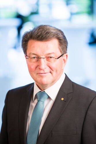 Älterer Geschäftsmann mit Brille und grauem Haar im dunklen Anzug und blau gestreifter Krawatte blickt lächelnd in die Kamera vor unscharfem, hellem Hintergrund