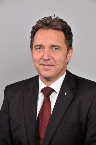 Mittelalterlicher Mann im dunklen Anzug mit weißem Hemd und bordeauxroter Krawatte lächelt vor neutral grauem Hintergrund in die Kamera