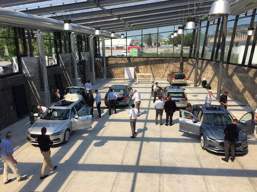 Menschen begutachten mehrere geparkte Limousinen in einer hellen, modernen Ausstellungshalle mit Glasdach und sichtbarer Belüftungsanlage