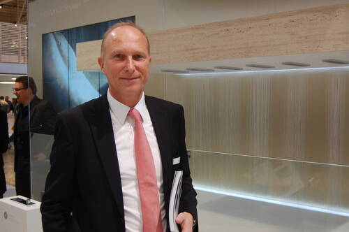 Mittelalter Mann im Anzug mit rosa Krawatte steht lächelnd vor einer modernen, horizontalen Duschinstallation auf einer Messe und hält Broschüren in der Hand.