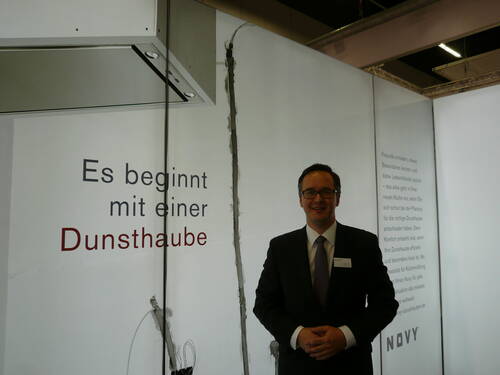 Mann im Anzug präsentiert auf einer Messe eine moderne Dunstabzugshaube vor einer Wand mit dem Slogan „Es beginnt mit einer Dunsthaube“