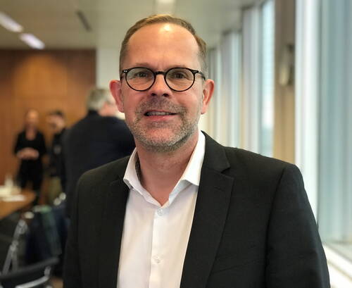 Mittelalterlicher Mann mit Brille im Anzug lächelt in einem modernen Konferenzraum mit unscharfen Gesprächsgruppen im Hintergrund