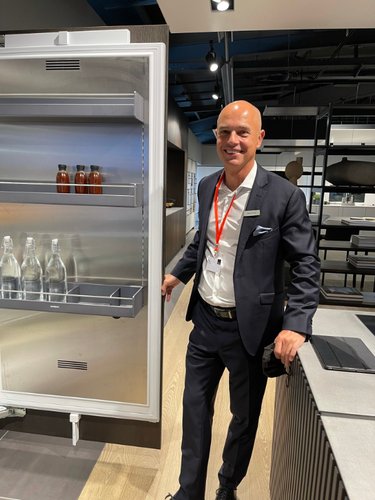 Eleganter Verkäufer im Anzug präsentiert einen geöffneten, modernen Einbaukühlschrank mit Getränkeflaschen in einem hochwertig eingerichteten Küchenschauraum