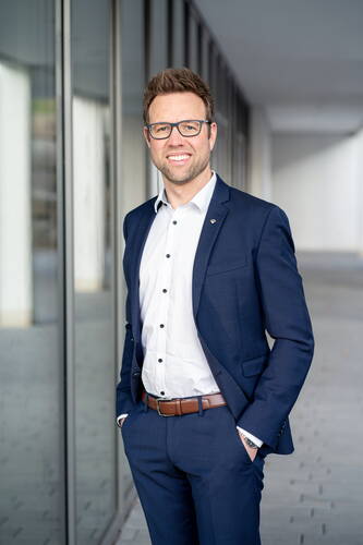 Lächelnder Mann im blauen Anzug mit Brille steht mit Händen in den Hosentaschen vor einer modernen Glasfassade in einem hellen Bürogebäude.
