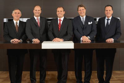 Fünf elegant gekleidete Geschäftsmänner stehen nebeneinander hinter einem modernen Konferenztisch und lächeln formell in die Kamera.