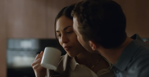 Mann küsst eine Frau zärtlich auf die Wange, während sie in der Küche aus einer weißen Tasse trinkt.