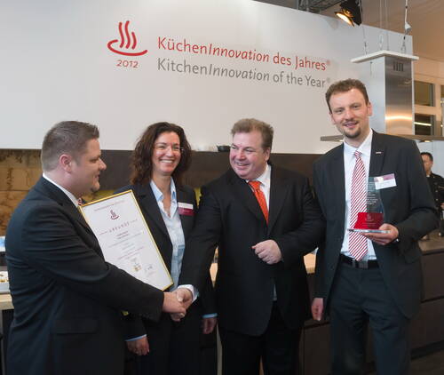 Vier Geschäftsleute in Anzügen feiern die Verleihung des Preises „KüchenInnovation des Jahres“, während einer eine Urkunde und ein anderer eine rote Trophäe hält und ein Handschlag erfolgt.