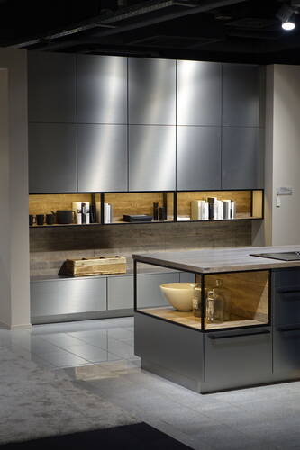 Modern gestaltete Küche mit matten Metallfronten, offen beleuchteten Holzregalen und Kücheninsel mit dekorativen Schalen und Flaschen
