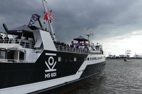 Passagierschiff MS KOI mit feiernden Menschen auf dem Oberdeck fährt bei dunklen Gewitterwolken durch einen Hafen, im Hintergrund weitere Schiffe und Hafenkräne