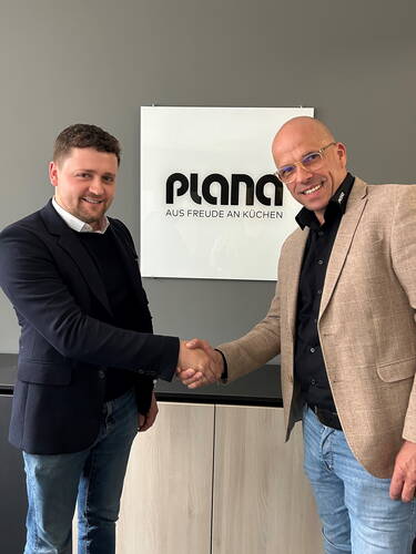 Zwei Geschäftsmänner in legerer Businesskleidung geben sich vor einem PLANA-Küchenstudio-Logo die Hand und lächeln in die Kamera