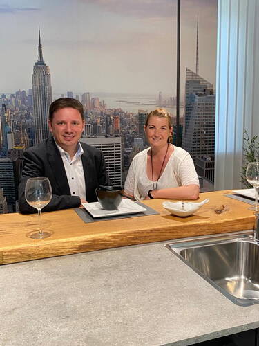 Zwei Personen sitzen an einer modern gedeckten Kücheninsel und lächeln in die Kamera, im Hintergrund eine großformatige Skyline von New York.
