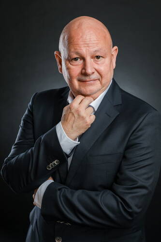 Glatzköpfiger Mann mittleren Alters im dunklen Business-Anzug blickt nachdenklich in die Kamera und stützt sein Kinn mit der Hand vor dunklem Hintergrund