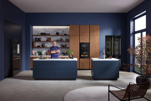 Mann in moderner, blau-goldener Designküche steht an Kochinsel mit Marmorplatte vor offenem Regal und hohen Holzfronten mit integrierten Backöfen
