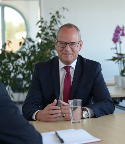 Geschäftsmann im Anzug mit roter Krawatte sitzt in einem modernen Büro am Konferenztisch, gestikuliert im Gespräch und hat ein Glas Wasser sowie Unterlagen vor sich.