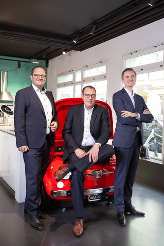 Drei Geschäftsleute in Anzügen posieren lächelnd in einem modernen Showroom vor einem knallroten Retro-Auto mit geöffnetem Kofferraum