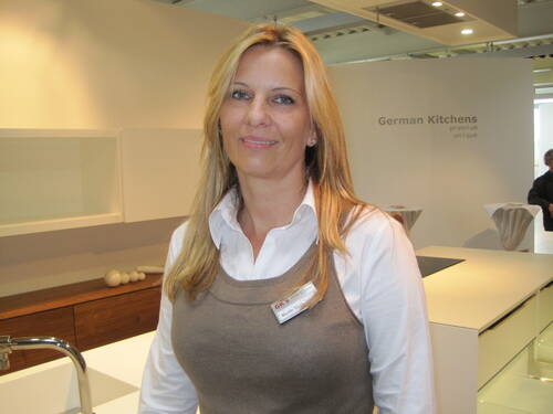 Blonde Verkaufsberaterin mit Namensschild steht lächelnd in einer modernen Ausstellungsküche mit hellen Arbeitsflächen und der Aufschrift „German Kitchens“ im Hintergrund