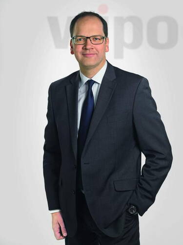 Geschäftsmann im dunklen Anzug mit Krawatte steht im Studio vor grauem Hintergrund und lächelt dezent in die Kamera.