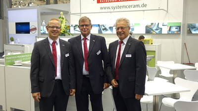Drei lächelnde Messemitarbeiter in dunklen Anzügen mit roten Krawatten posieren vor einem Systemceram‑Messestand mit weißen Tischen und Küchenexponaten.